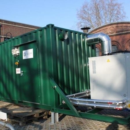 Grüner Schiffscontainer mit externen Maschinen, der im Freien in der Nähe eines Backsteingebäudes steht