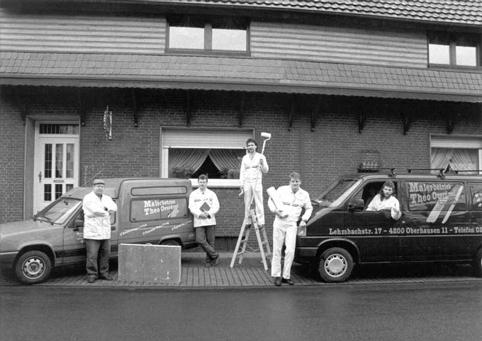 Fünf Personen stehen neben einem Lieferwagen und einem Lastwagen vor einem Gebäude, möglicherweise eine Arbeitsgruppe.